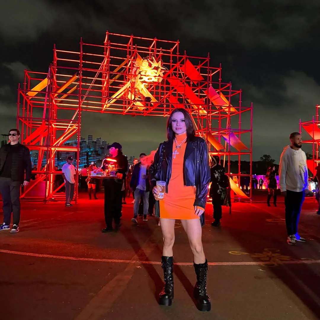 Lahra de jaqueta preta, vestido laranja e botas diante de uma grande estrutura vermelha, à noite