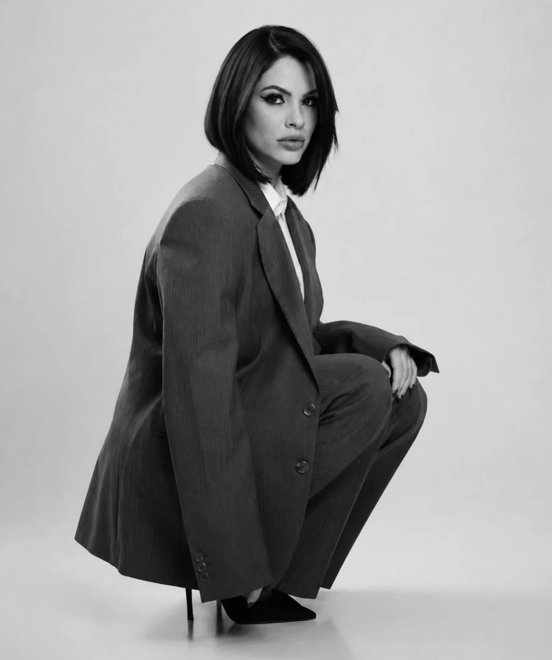 Lahra em preto e branco, agachada com um terno oversized e cabelo curto
